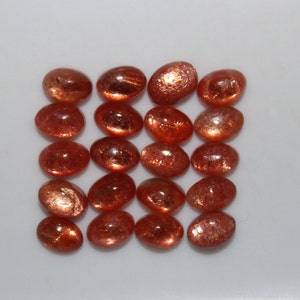 May include: A collection of 18 oval-shaped sunstone gemstones. The stones are a mix of orange and red hues with a shimmery, iridescent finish.