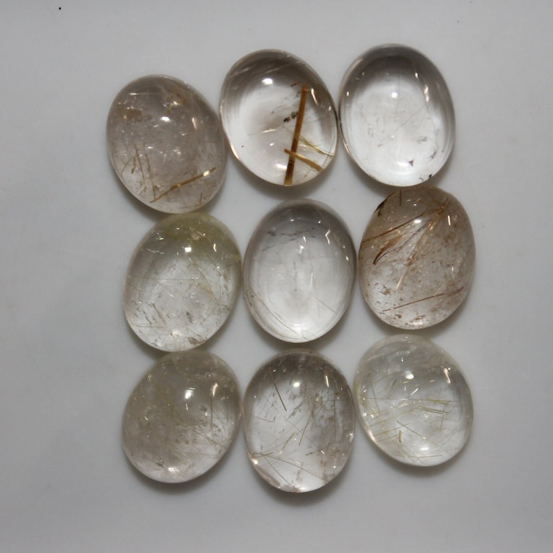May include: Ten oval-shaped, clear quartz cabochons with gold rutile inclusions. The cabochons are smooth and polished.