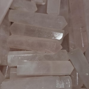 May include: A collection of pale pink quartz crystal wands. Each wand is rectangular with a pointed end. The crystals are arranged closely together, filling the frame. The lighting creates highlights and shadows on the surfaces of the crystals.
