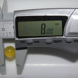 May include: A digital caliper measuring a yellow sphere. The digital display shows 8.00 mm. The caliper is silver and has buttons for mm, inch, on/off, and zero. The sphere is approximately 0.31 inches in diameter.