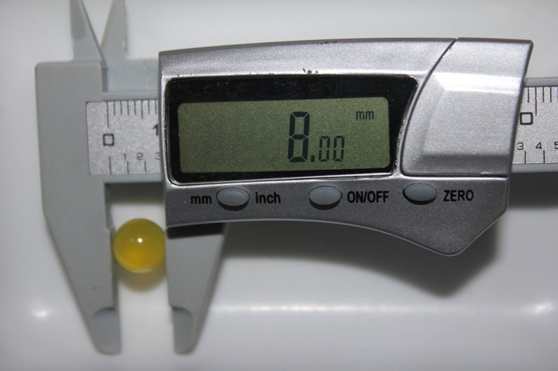 May include: A digital caliper measuring a yellow sphere. The digital display shows 8.00 mm. The caliper is silver and has buttons for mm, inch, on/off, and zero. The sphere is approximately 0.31 inches in diameter.