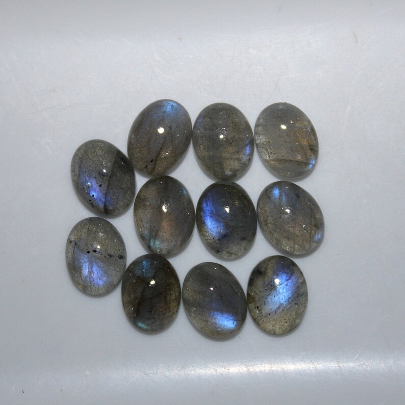 May include: Ten oval-shaped labradorite gemstones with a blue iridescent sheen. The stones are arranged in a cluster on a white surface.