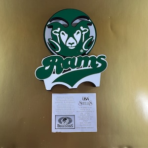 Puede incluir: Un recorte de espuma 3D verde y blanco de una cabeza de carnero con la palabra "Rams" en letras verdes debajo. El recorte está hecho por Shelia's, una empresa que crea diseños personalizados para eventos corporativos, recaudación de fondos locales y promociones escolares. La información de contacto de la empresa se incluye en la parte inferior del recorte.