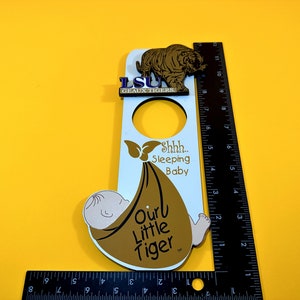 Può includere: Appendiporta bianco con un tigre marrone e un bambino che dorme in una coperta marrone. L'appendiporta ha un foro al centro e dice "LSU Geaux Tigers" e "Shhh... Sleeping Baby Our Little Tiger".