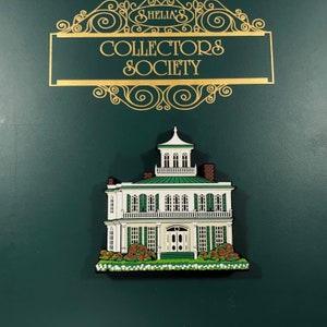 May include: A white and green painted wooden pin of a two-storey house with a green roof and white trim. The house has a porch with white railings and a front door with a green door frame. The house is surrounded by green bushes and white flowers. The pin is on a green background with a gold frame that says "Shellia's Collectors Society".