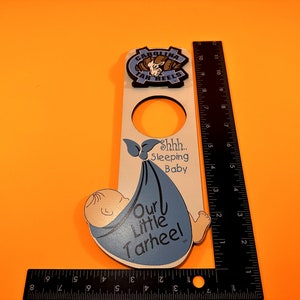 以下が含まれることがあります： 青い毛布にくるまれた青い赤ちゃんが描かれた白い木製のドアハンガー。赤ちゃんは寝ていて、ハンガーには「Shhh... 寝ている赤ちゃん 私たちの小さなTarheel」と書かれています。ハンガーの上部には、カロライナ・ターヒールズのロゴも付いています。