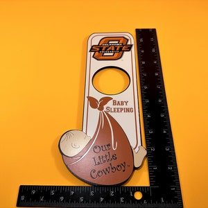 May include: A wooden door hanger with the Oklahoma State University logo and the words "Baby Sleeping" and "Our Little Cowboy." The hanger is approximately 10 inches long and has a cutout for the doorknob.