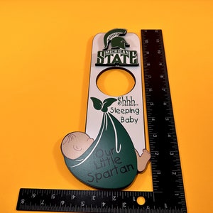 Puede incluir: Un colgador de puerta de madera blanco con un bebé verde envuelto en una manta verde. El bebé tiene una cara marrón claro y está durmiendo. El texto "Shhh... Sleeping Baby" está escrito en negro en el colgador. El texto "Our Little Spartan" está escrito en verde en el colgador. El logotipo del Spartan del estado de Michigan está en la parte superior del colgador.