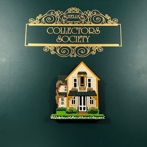 May include: A yellow and green painted wooden house with a black roof and white trim. The house has a porch with a railing and a door with a screen. The house is surrounded by green bushes and flowers. The house is part of a collector's society.