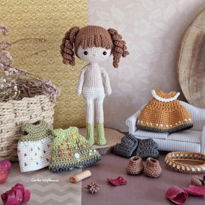 Pode incluir: Boneca de crochê com cabelo castanho e um vestido branco, juntamente com três vestidos de crochê em cores diferentes e uma faixa de cabeça de crochê. A boneca está sentada em uma superfície de madeira com um pequeno sofá atrás.