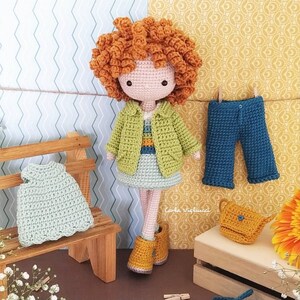 May include: Crocheted doll with curly orange hair wearing a green and yellow jacket and a light blue skirt. The doll is holding a small yellow purse. The doll is sitting on a wooden bench with a light green crocheted dress on the bench. There is a pair of blue crocheted pants hanging on a clothesline. The background is a light blue and yellow patterned wall.