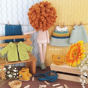 May include: A crocheted doll with red hair, wearing a variety of crocheted clothing, including a green jacket, blue jeans, and two dresses. The doll is sitting on a wooden bench with a small flower arrangement and a crocheted bag.