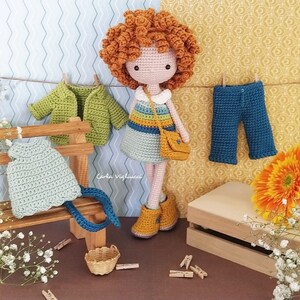 May include: Crochet doll with red hair wearing a blue and yellow dress and a brown bag. The doll is standing on a wooden bench with a green jacket, a blue dress, and a pair of blue pants hanging on it. The doll is surrounded by flowers and a wooden box.