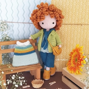 May include: Crocheted doll with red curly hair wearing a green jacket, blue jeans, and brown shoes. The doll is holding a small brown bag. The doll is sitting on a wooden bench with a crocheted dress on the bench. The background is a yellow and white patterned wall.