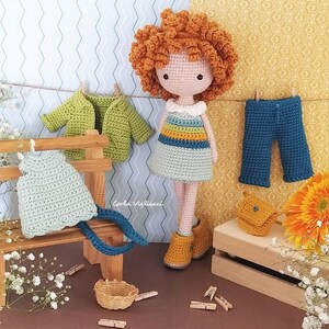 May include: A crocheted doll with red curly hair wearing a striped dress and brown boots. The doll is surrounded by crocheted clothing items, including a green cardigan, a blue pair of pants, and a light blue dress. The doll is sitting on a wooden bench with a basket and clothespins.