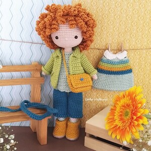 May include: Crocheted doll with curly red hair wearing a green jacket, blue jeans, and a yellow bag. The doll is holding a small crocheted bag. The doll is sitting on a wooden bench with a crocheted dress hanging on a clothesline behind it.
