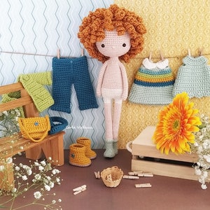 May include: A crocheted doll with curly orange hair wearing a white crocheted dress and white underwear. The doll is standing on a wooden surface with a small wooden bench, a crocheted bag, and a pair of yellow crocheted boots. There are also several crocheted clothes hanging on a clothesline.