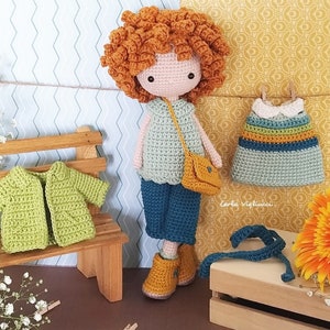 May include: Crochet doll with red curly hair wearing a blue and green outfit, a yellow bag, and brown shoes. The doll is holding a blue crochet strap. The doll is sitting on a wooden bench with a green crochet jacket and a blue crochet dress hanging on a wooden crate.
