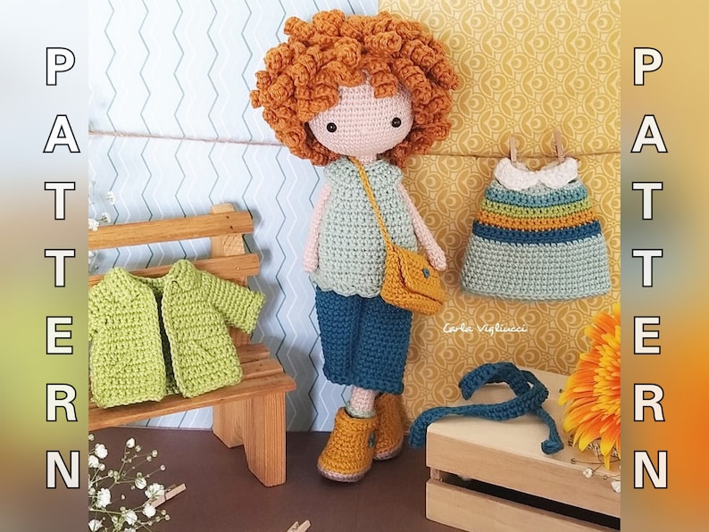 May include: Crochet doll with red curly hair wearing a blue and green outfit, a yellow bag, and brown shoes. The doll is holding a blue crochet strap. The doll is sitting on a wooden bench with a green crochet jacket and a blue crochet dress hanging on a wooden crate.