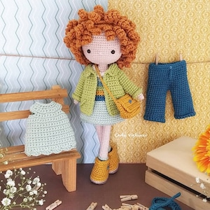 May include: A crocheted doll with brown curly hair wearing a green jacket, a yellow bag, and a blue and white dress. The doll is standing on a wooden bench with a green crocheted dress on it. The background is a patterned wall with the word "PATTERN" on either side.