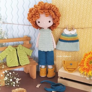 May include: Crochet doll with curly red hair wearing a green sweater, blue pants, and yellow boots. The doll is holding a blue and yellow striped dress and a yellow purse. The doll is sitting on a wooden bench with a green crocheted jacket on top.