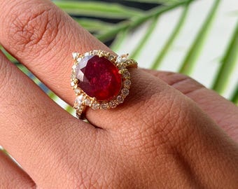 May include: A gold ring featuring a large, oval-cut deep red gemstone at the centre, surrounded by a halo of small, clear, round-cut stones. The band is thin and also set with small, clear, round-cut stones along the visible shank.