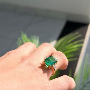 May include: A gold ring featuring a large, rectangular, emerald-cut green gemstone as the centrepiece, flanked by a smaller, round, clear gemstone on one side, set on a thin, polished gold band that encircles the finger.