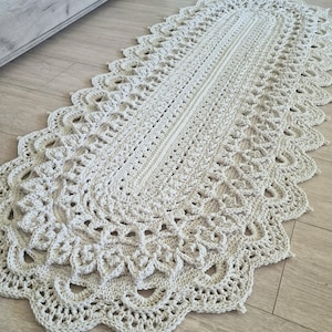 Könnte beinhalten: Ein gehäkelter ovaler Teppich mit einem zarten, filigranen Muster in Creme. Der Teppich hat einen gewellten Rand und ist aus dickem Garn gefertigt.