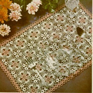 May include: A white crocheted table runner with a delicate floral pattern and a pink border. The runner is laid out on a table with a glass and silverware.