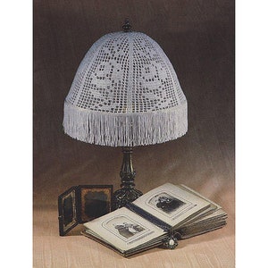 May include: A white crocheted lampshade with a fringe trim sits on a table lamp with a dark brown base. The lamp is next to a closed book with a vintage photo on the cover.