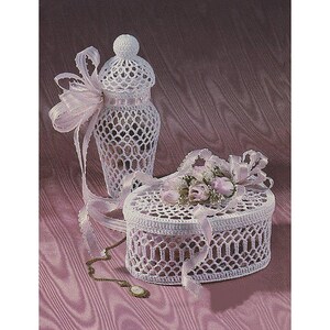 May include: A white, crocheted decorative vase with a matching lid and a round, lidded box. Both items are adorned with delicate pink ribbons and small pink roses. A gold-toned necklace with a cameo pendant rests near the box.