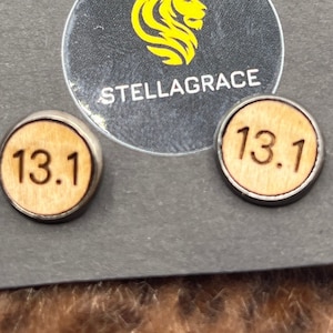 May include: A pair of wooden earrings with a silver metal backing. The earrings are round and feature the number "13.1" engraved on them. The earrings are displayed on a black card with the text "Stellagrace" printed on it.