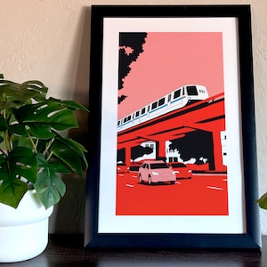 May include: A framed print of a stylized illustration of a train traveling over a red overpass with cars driving on the road below. The background is a pink and red gradient.