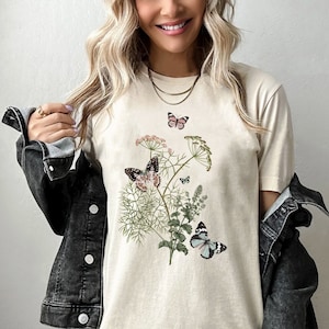 May include: A beige t-shirt with a floral design featuring butterflies. The butterflies are in shades of pink, black, and blue. The design is centered on the shirt.