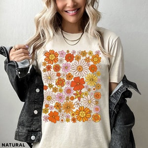May include: A beige t-shirt with a retro floral print featuring orange, yellow, pink, and white flowers. The shirt has a crew neck and short sleeves.