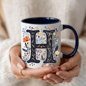 May include: White ceramic mug with a navy blue interior and handle. The mug features a floral design with a navy blue letter "H" in the center. The floral elements include orange, yellow, and blue flowers and green leaves.