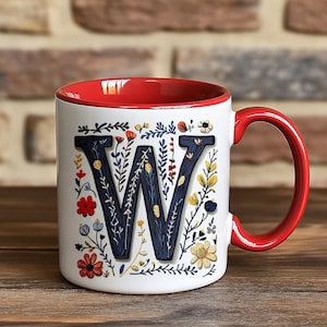 May include: White ceramic mug with a red rim and handle. The mug features a large, stylized letter "W" surrounded by colorful floral designs.