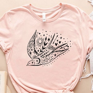 May include: A pink t-shirt featuring a black graphic of a stylised bird with floral and star accents.