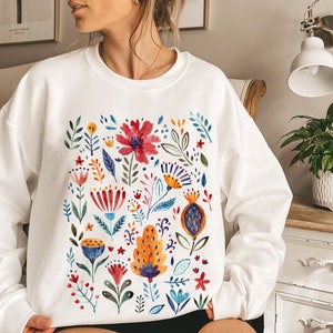 May include: White sweatshirt with a colorful floral print. The flowers are painted in a watercolor style and include red, yellow, blue, green, and orange hues.