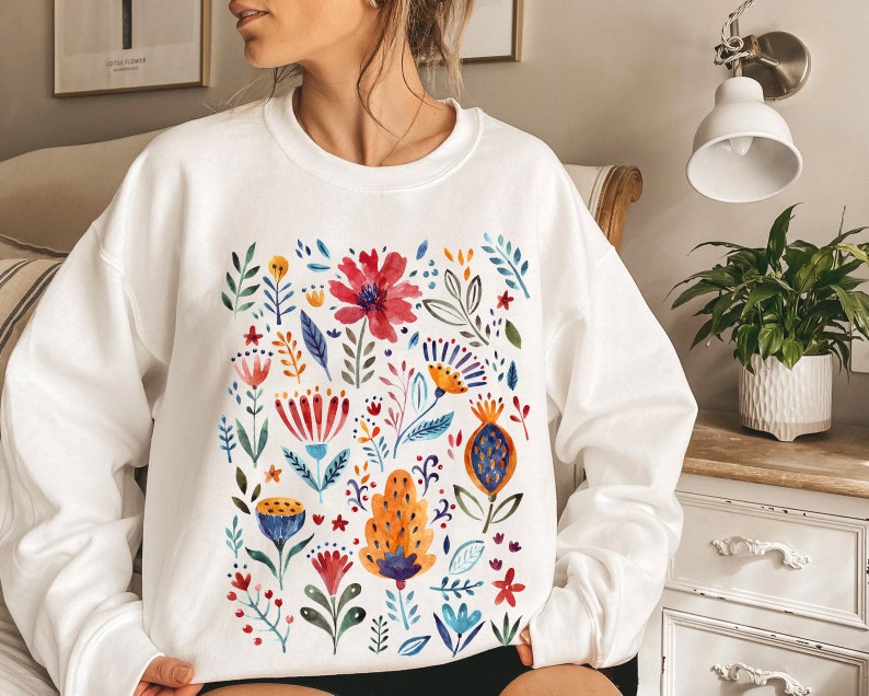 May include: White sweatshirt with a colorful floral print. The flowers are painted in a watercolor style and include red, yellow, blue, green, and orange hues.