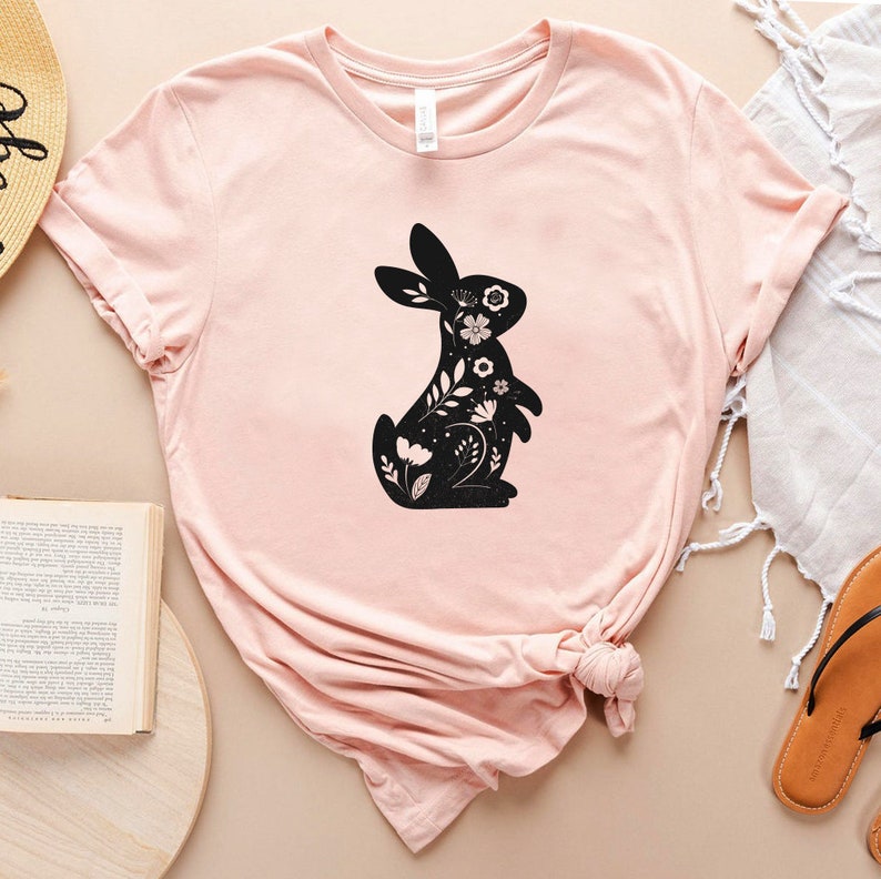 K&ouml;nnte beinhalten: Ein pinkfarbenes T-Shirt mit einer schwarzen Silhouette eines Kaninchens mit floralen Details. Das Kaninchen blickt nach links.