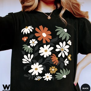 May include: Black t-shirt featuring a floral design with white, orange, and green flowers. The graphic includes dark gray leaves and stems. The shirt has a crew neck and short sleeves. The word "BLACK" is in a circle on the bottom right.