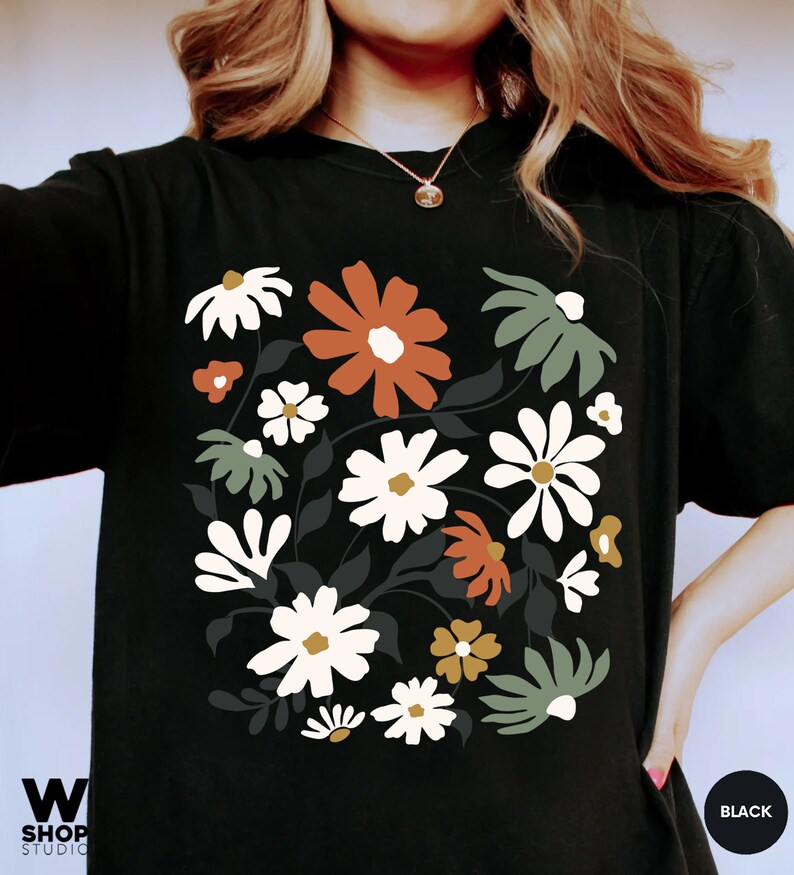 May include: Black t-shirt featuring a floral design with white, orange, and green flowers. The graphic includes dark gray leaves and stems. The shirt has a crew neck and short sleeves. The word "BLACK" is in a circle on the bottom right.