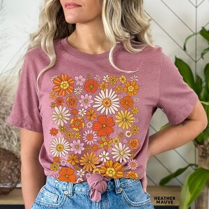 May include: A mauve t-shirt with a retro floral print featuring orange, yellow, and pink flowers. The shirt is tied at the waist and the text "HEATHER MAUVE" is visible at the bottom of the image.