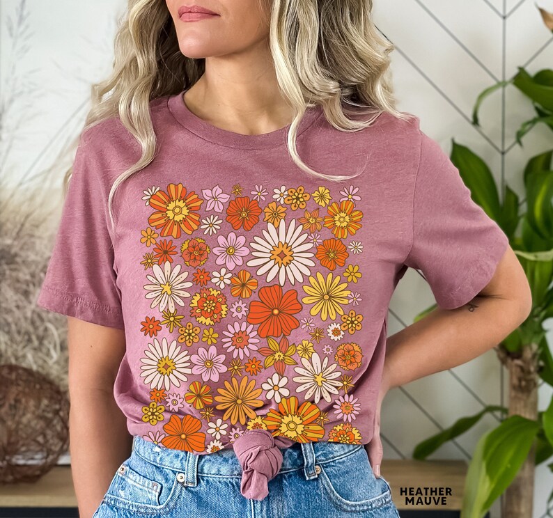 May include: A mauve t-shirt with a retro floral print featuring orange, yellow, and pink flowers. The shirt is tied at the waist and the text "HEATHER MAUVE" is visible at the bottom of the image.