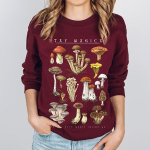 Op de afbeelding: Een bordeauxrood sweatshirt met een paddenstoelprint en de tekst "STAY MAGICAL KEEP MAGIC INSIDE US".
