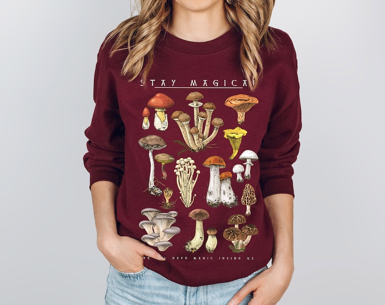 Op de afbeelding: Een bordeauxrood sweatshirt met een paddenstoelprint en de tekst "STAY MAGICAL KEEP MAGIC INSIDE US".