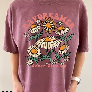 Puede incluir: Una camiseta morada con un gr&aacute;fico de flores y el texto "Daydreamer Never Give Up".