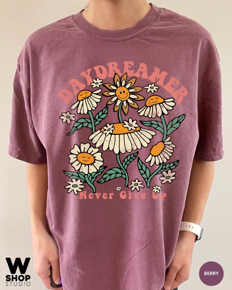 Puede incluir: Una camiseta morada con un gr&aacute;fico de flores y el texto "Daydreamer Never Give Up".