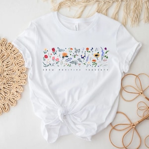 May include: White t-shirt with a floral design and the text "Grow Positive Thoughts".